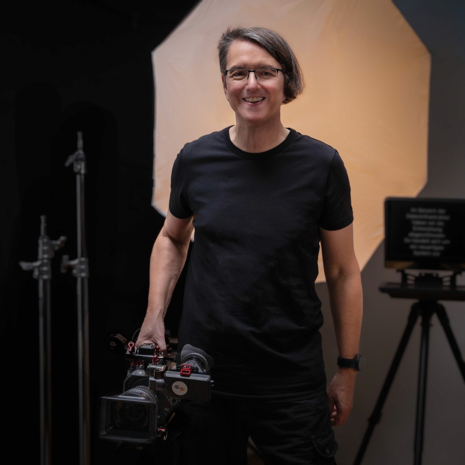 Alex Mikula am Set mit einer Kamera in der Hand und einem Teleprompter und einem Scheinwerfer im Hintergrund