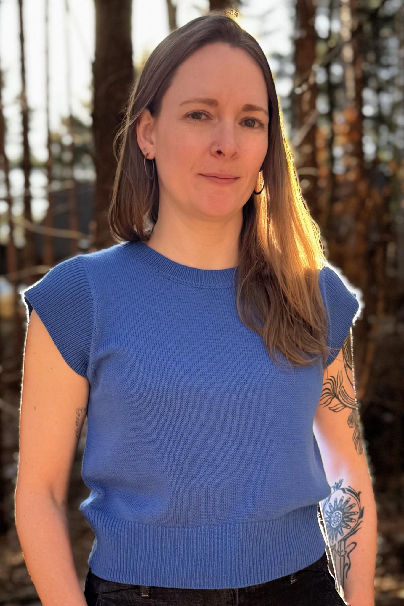 Woman in a blue shirt with long brown hair, standing  with a forest in the background