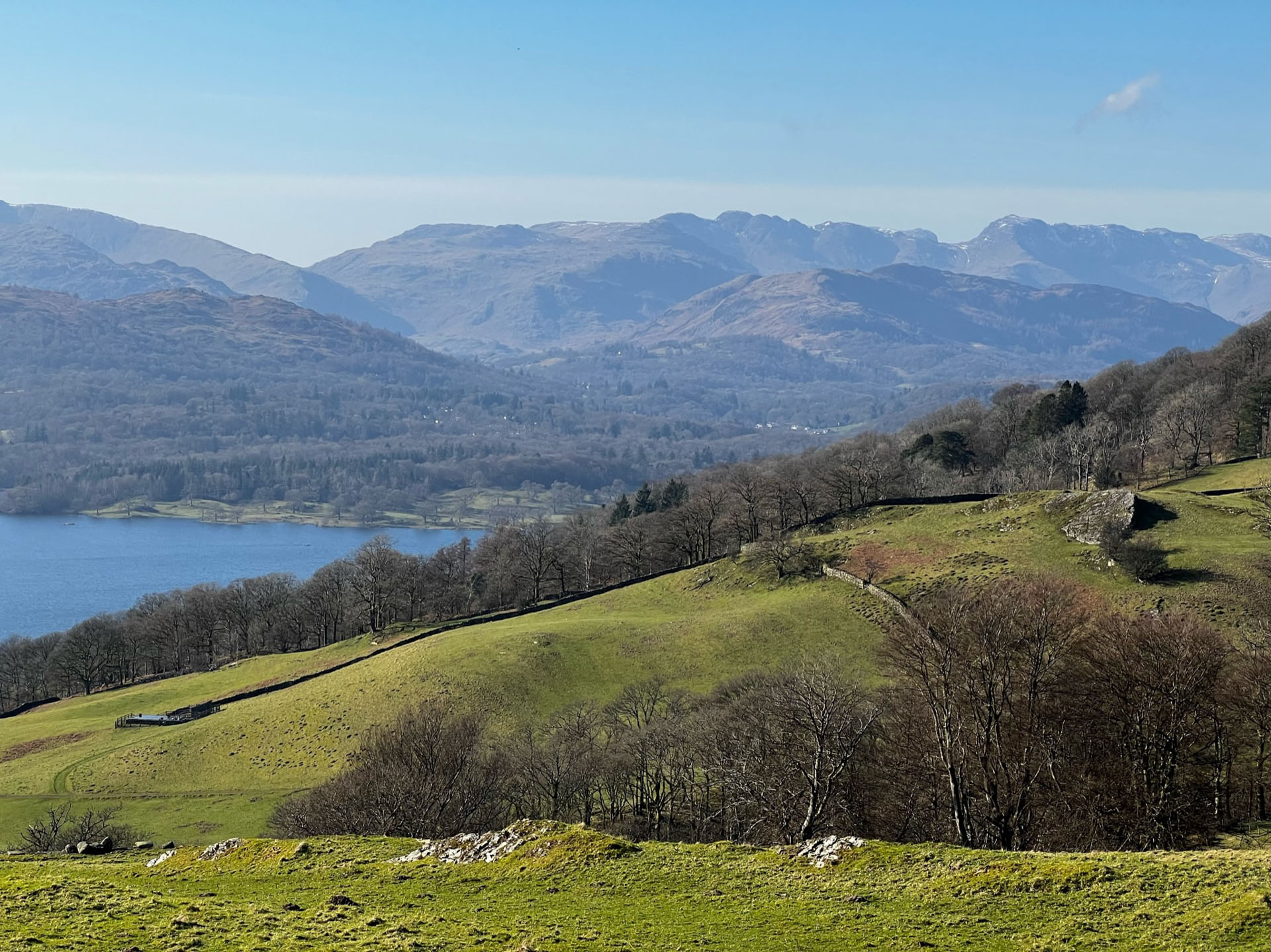 Sloping hills lead down into Windermere lake. Fells are seen in the background, still with a little snow on the top. Clear blue skies.