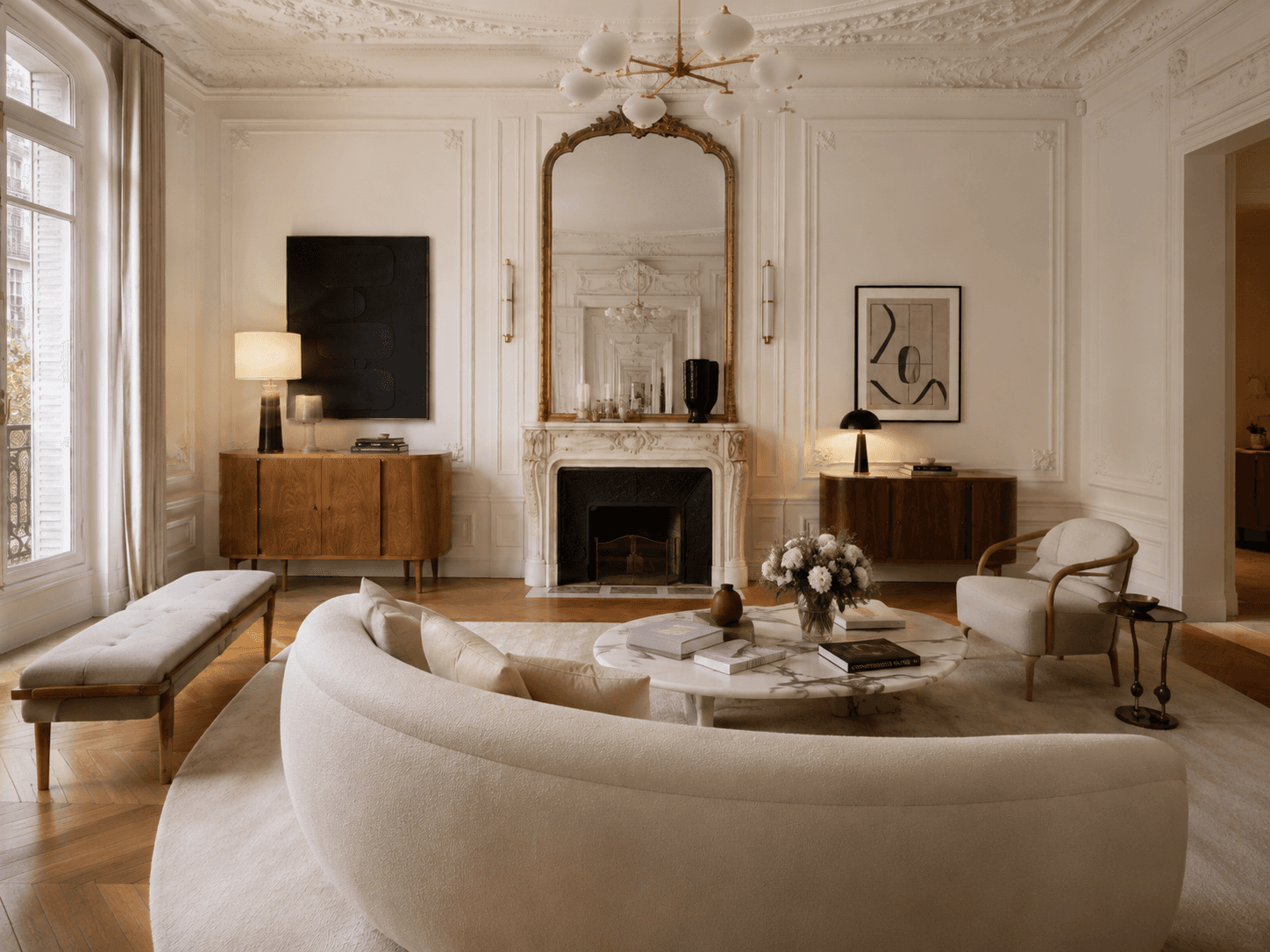 Refined Haussmann apartment living room from behind the curved sofa, showing a marble fireplace, gilded mirror, abstract art, and warm wood accents.
