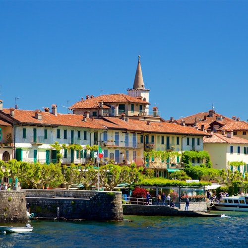 Colorful waterfront buildings with red roofs, green shutters, and lush plants, beside a lake under a clear blue sky.