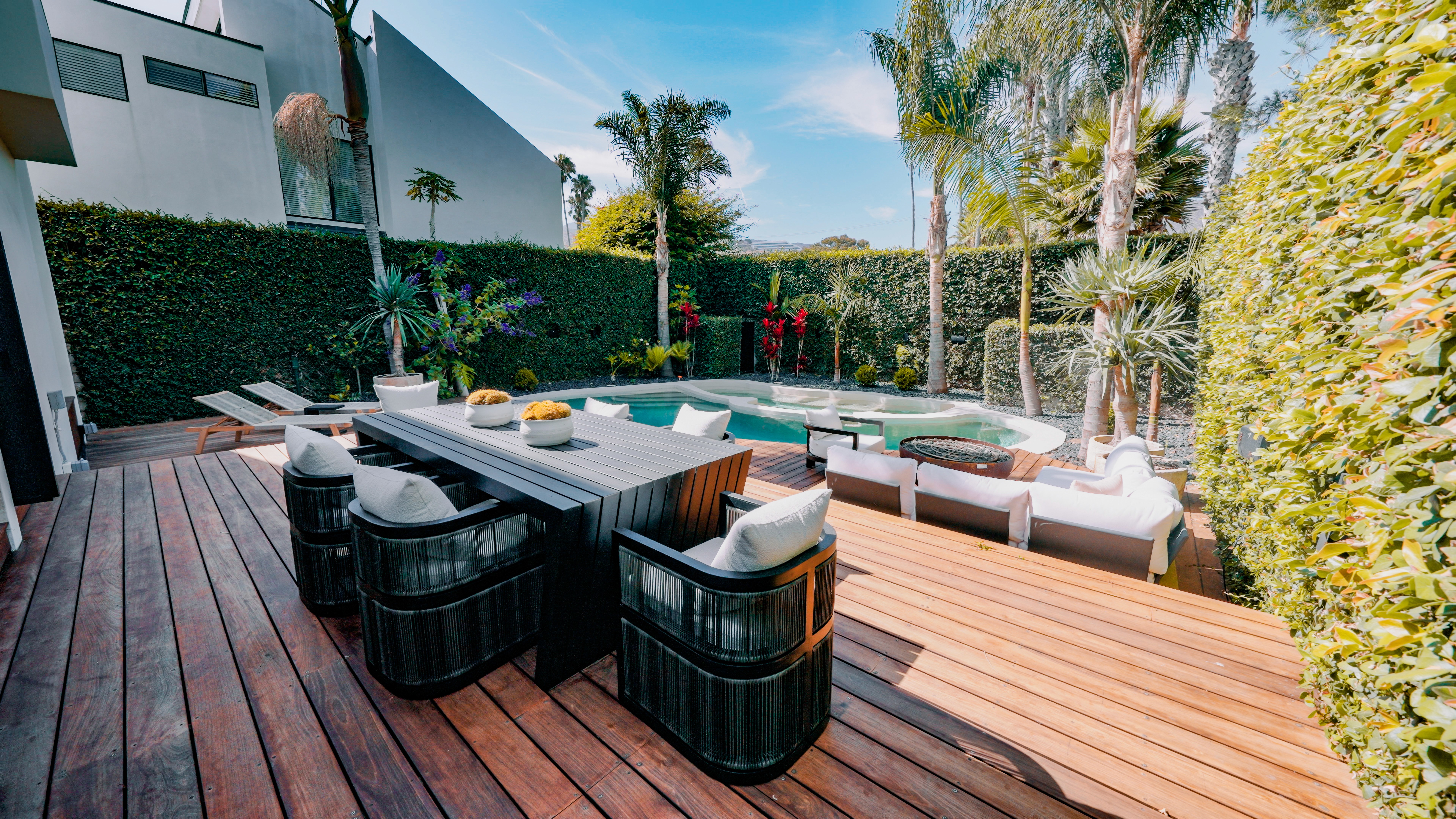 Outdoor dining set on the deck at Tideline Malibu Colony with pool, fire pit, and lush privacy hedge in the background