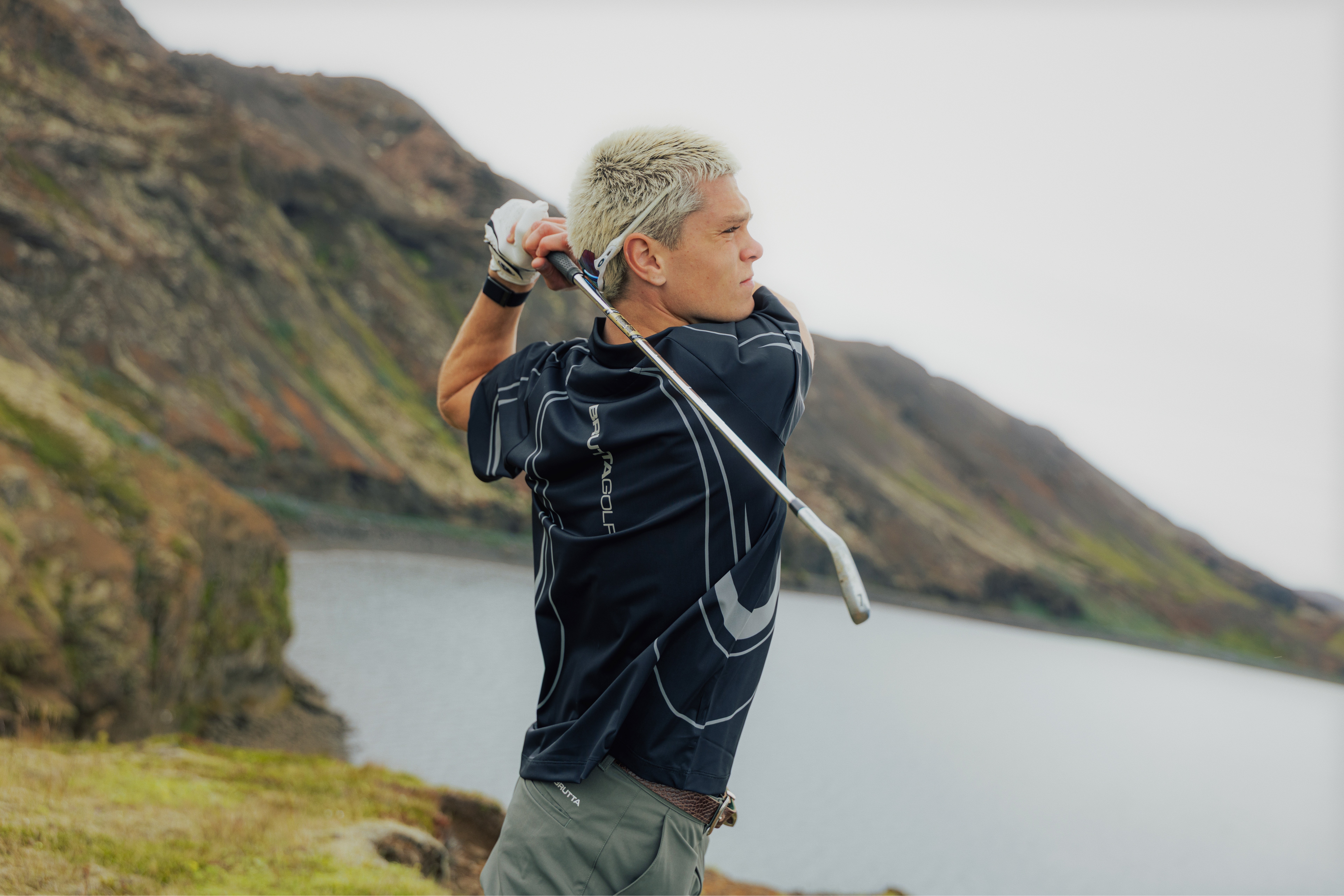 Golfer following through on a drive beside a lake, Icelandic hills behind