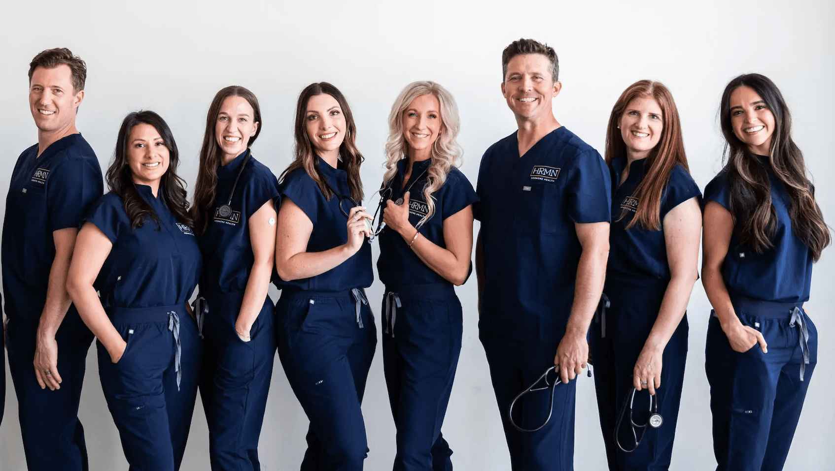Group photo of the HRMN Health team in matching blue scrubs, standing in front of shelves stocked with medical products, smiling in a professional and welcoming clinical setting.