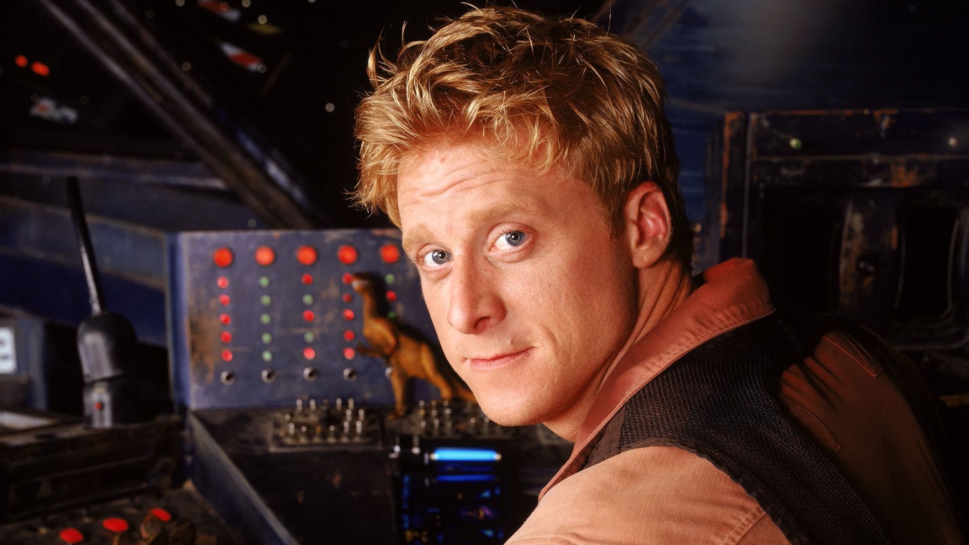 A person with short blonde hair wearing a brown shirt and dark vest is sitting in a dimly lit cockpit, looking over their shoulder. Various control panels with lights and switches are visible in the background.