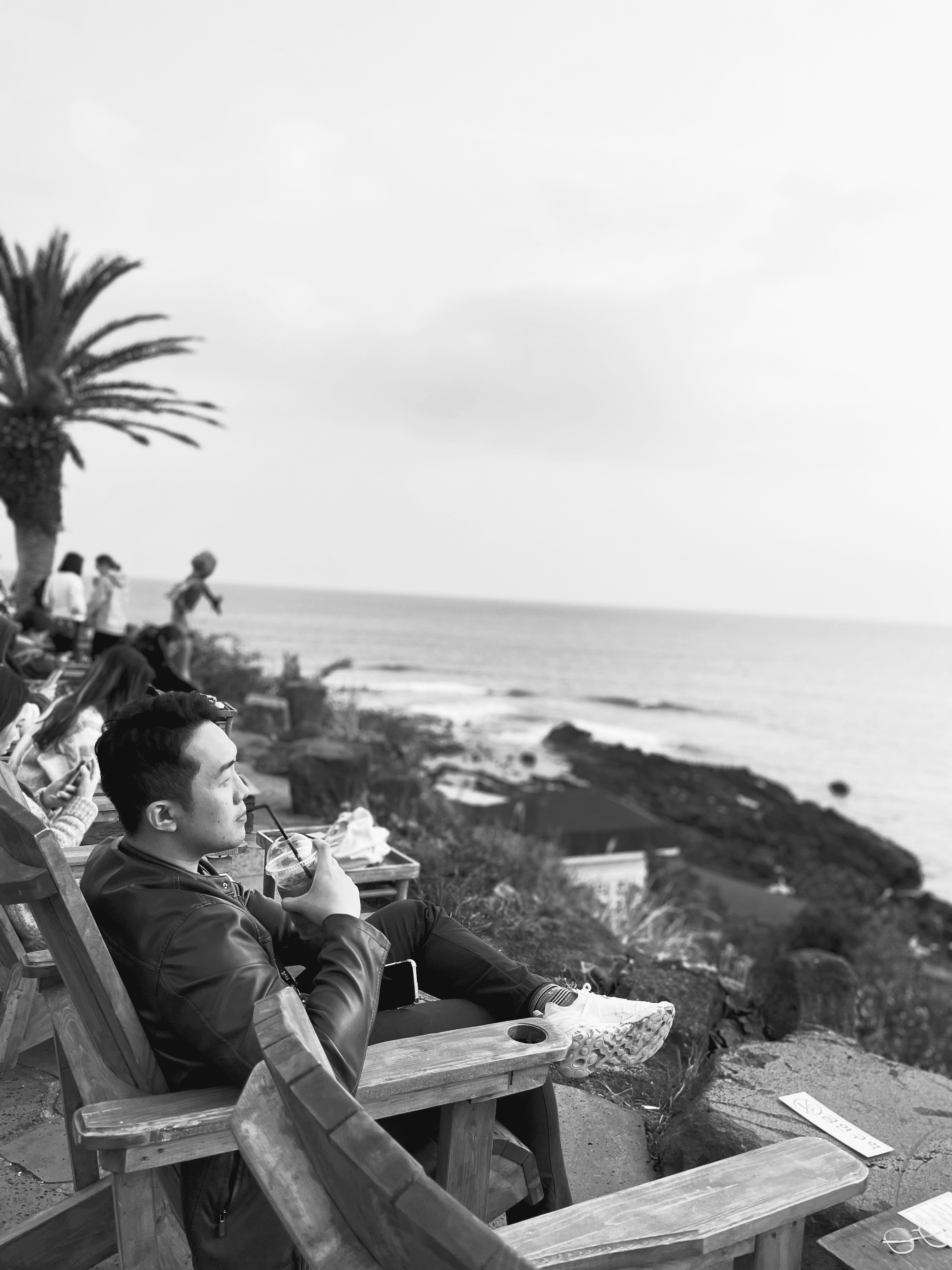 A black and white photo of an ocean view with a palm tree and people relaxing on wooden deck chairs.