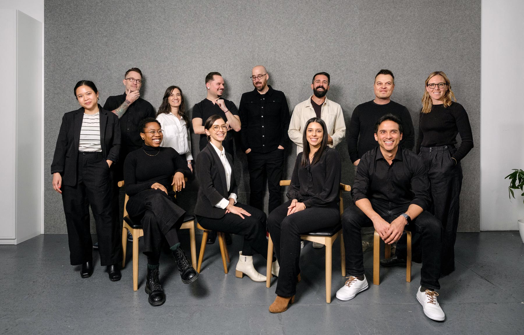 Professional studio portrait of The UP Studio architecture team, featuring eleven members dressed in coordinated black and white attire. Some team members are seated on wooden chairs while others stand behind them against a grey backdrop. The composition shows a mix of casual and professional styling, with varied footwear choices adding personality to the unified look.