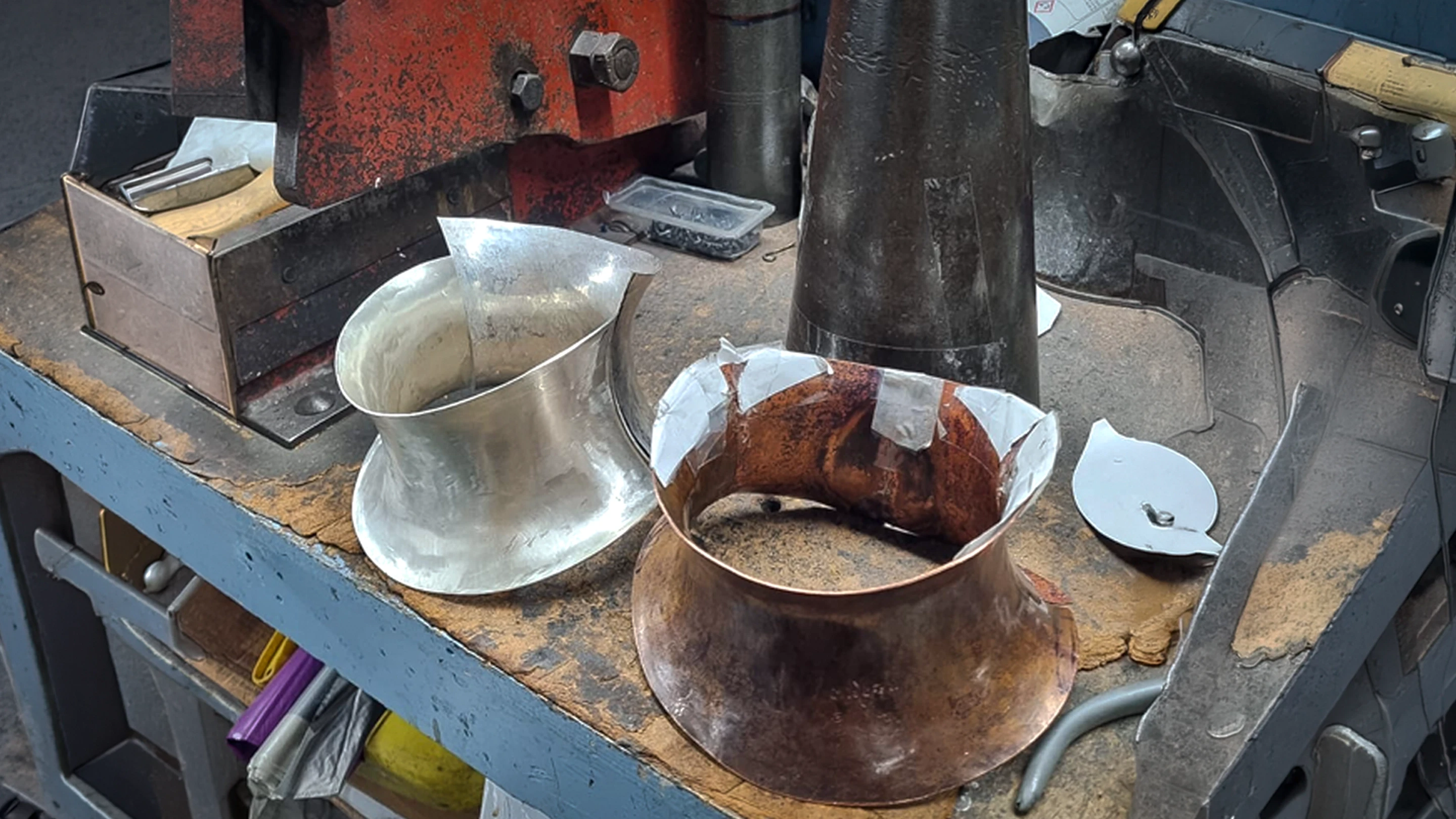 A silver and copper neck collar prototype shaped around a metal mandrel on a silversmithing workbench, surrounded by workshop tools