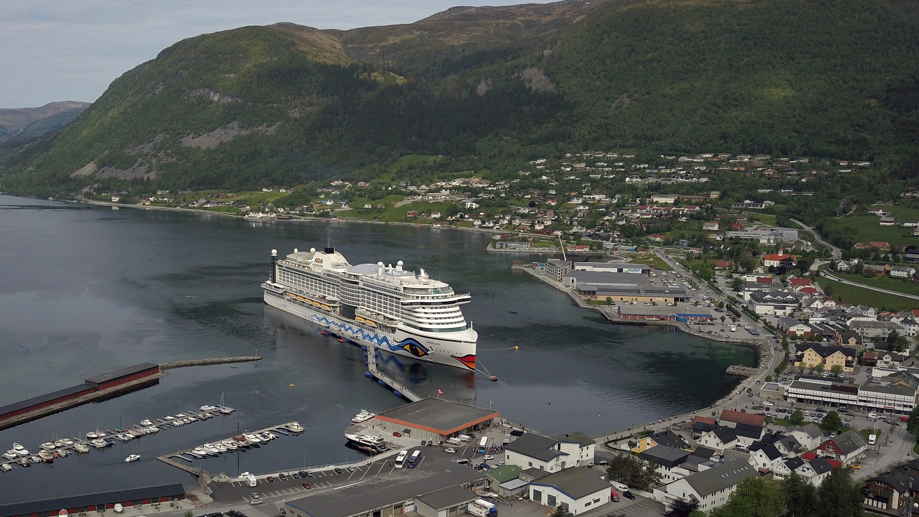 Aerial view of a cruise ship moored to a floating pier in Nordfjordeid, with shoreline buildings and surrounding mountains in the background.