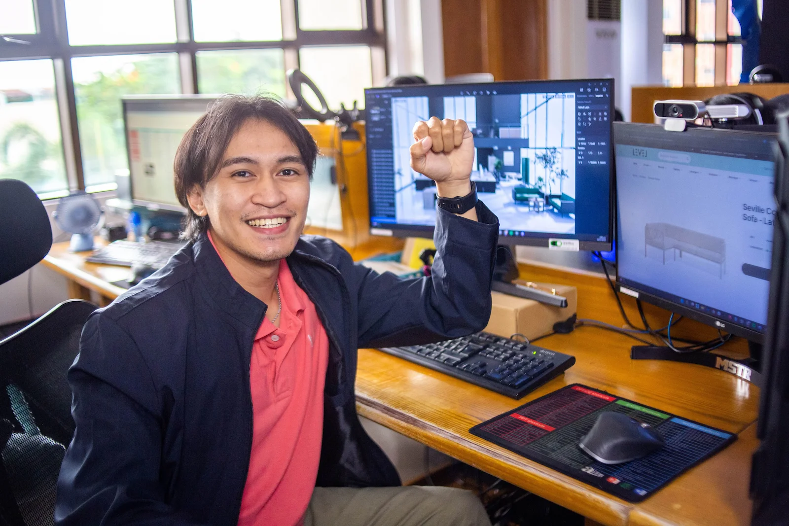 A smiling man gestures energetically while sitting at a dual-monitor workstation in a creative office.