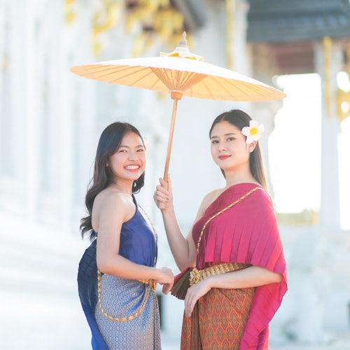 两名穿着传统服饰的女性站在户外，一人手持油纸伞。两人都面带微笑，背景中有装饰性的建筑物。