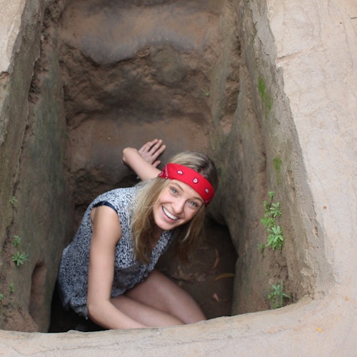Seorang pria yang mengenakan bandana merah dan atasan abu-abu berjongkok dan tersenyum di dalam parit sempit berbatu yang dikelilingi oleh tanah dan tanaman kecil.