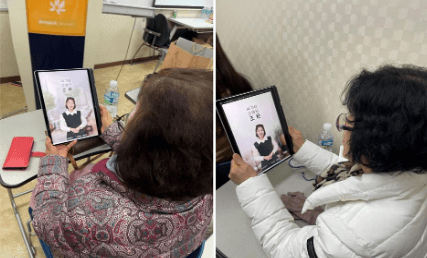Two women holding tablets, each displaying a person on the screen. The tablets appear to be in a virtual meeting setting.