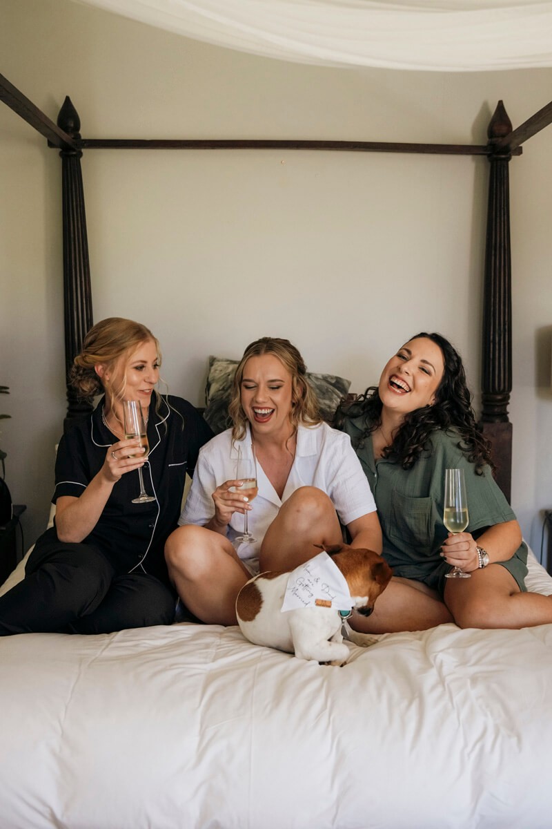 Bride and bridesmaids laughing on bed during wedding morning preparations