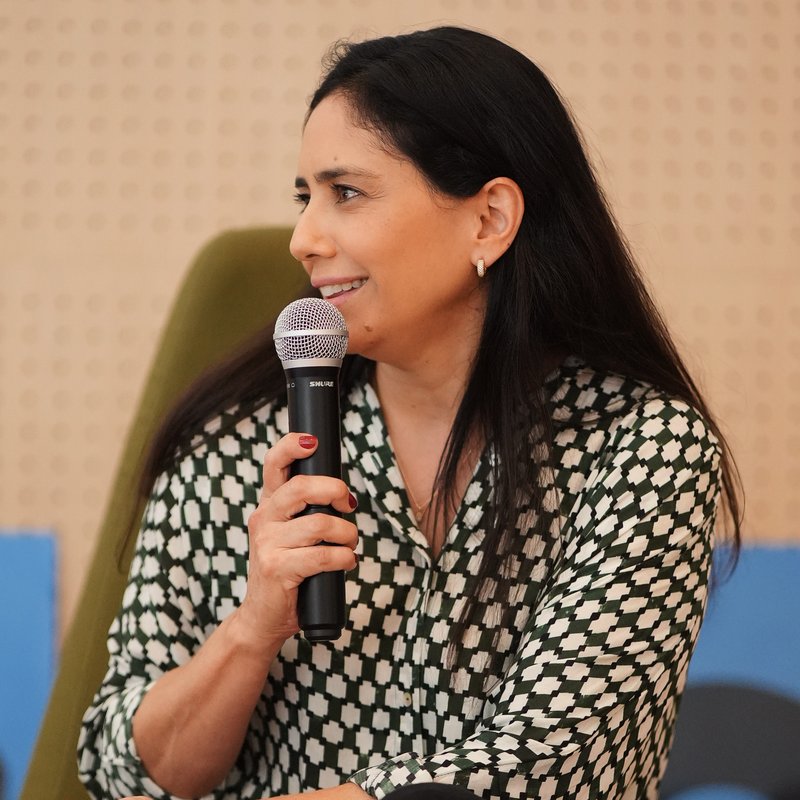 Alessandra Caribé falando ao microfone em evento, de blusa estampada verde e branco.