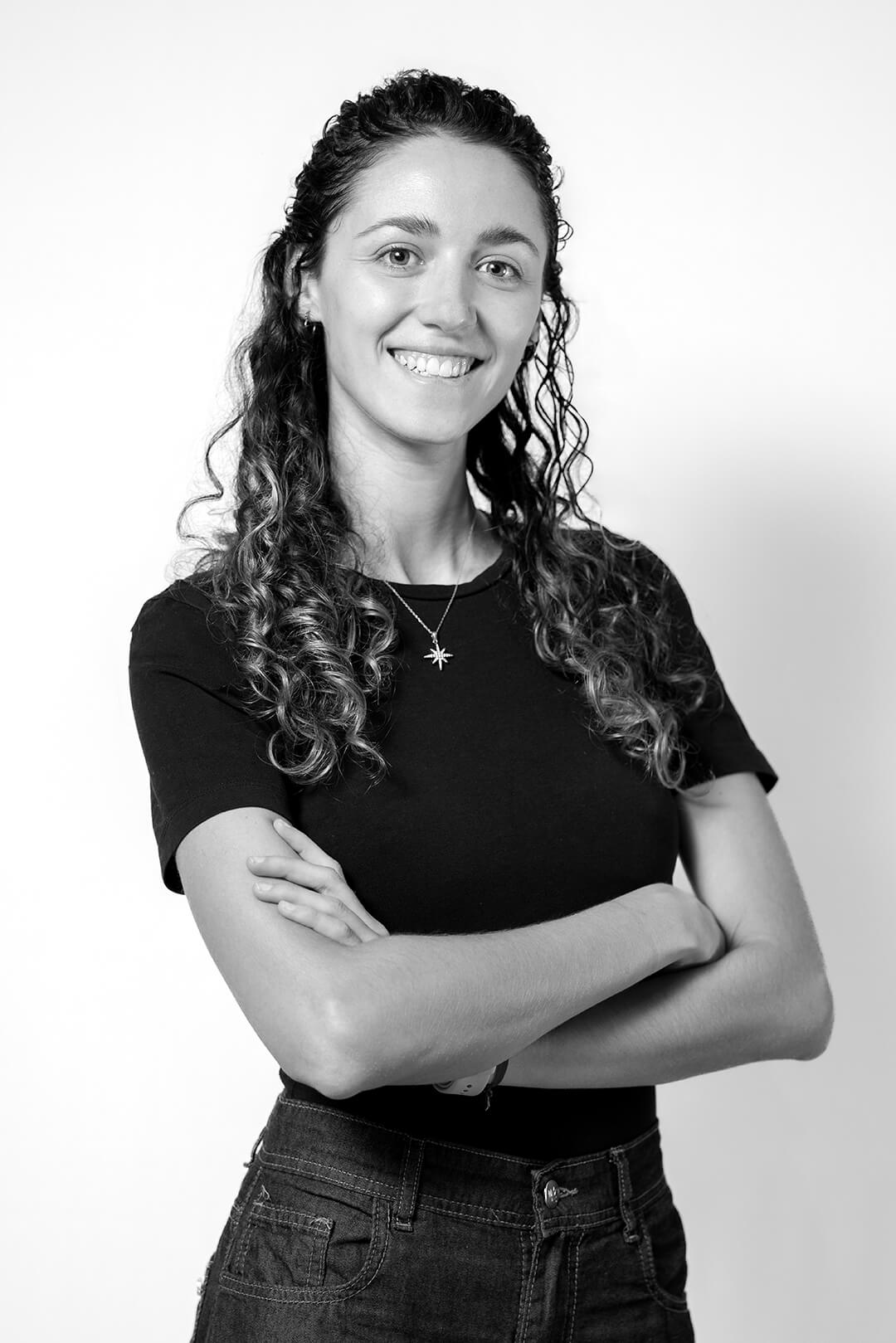 Smiling woman with curly hair, wearing a black t-shirt and necklace, stands confidently with arms crossed against a plain white background.