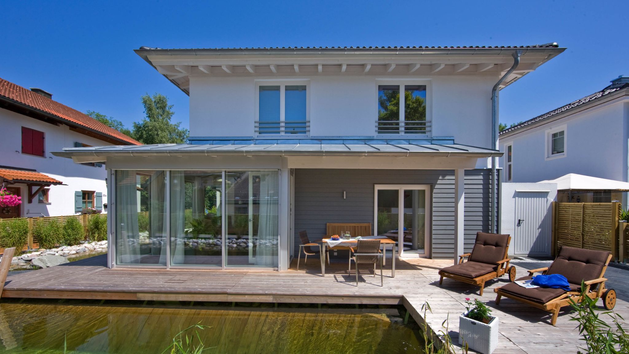 Moderne Villa mit Holzdeck, Teich, Gartenmöbeln und Wintergarten an einem sonnigen Tag.