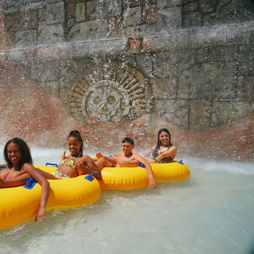 Four people in yellow inflatable tubes float under a waterfall with a stone wall featuring a decorative carving in the background.