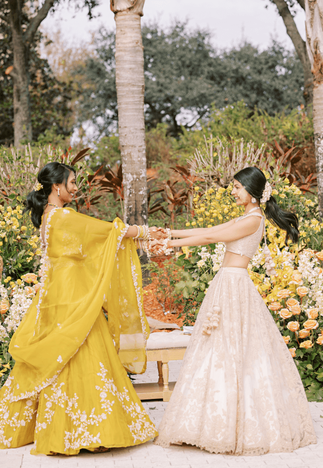 Two women in traditional Indian lehengas holding hands and smiling at a wedding ceremony