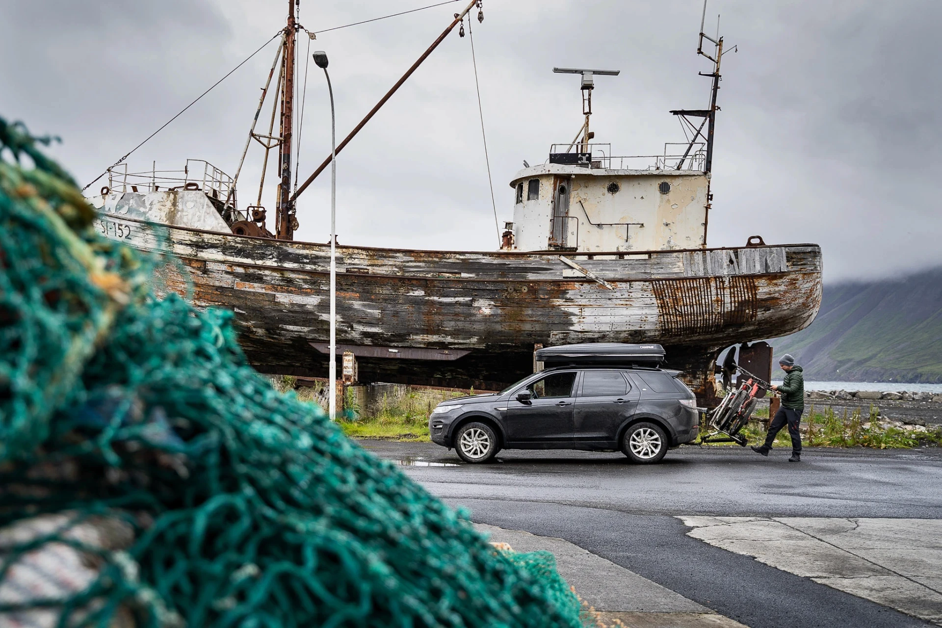 Car with bikes parked beside an old fishing boat on a grey day.