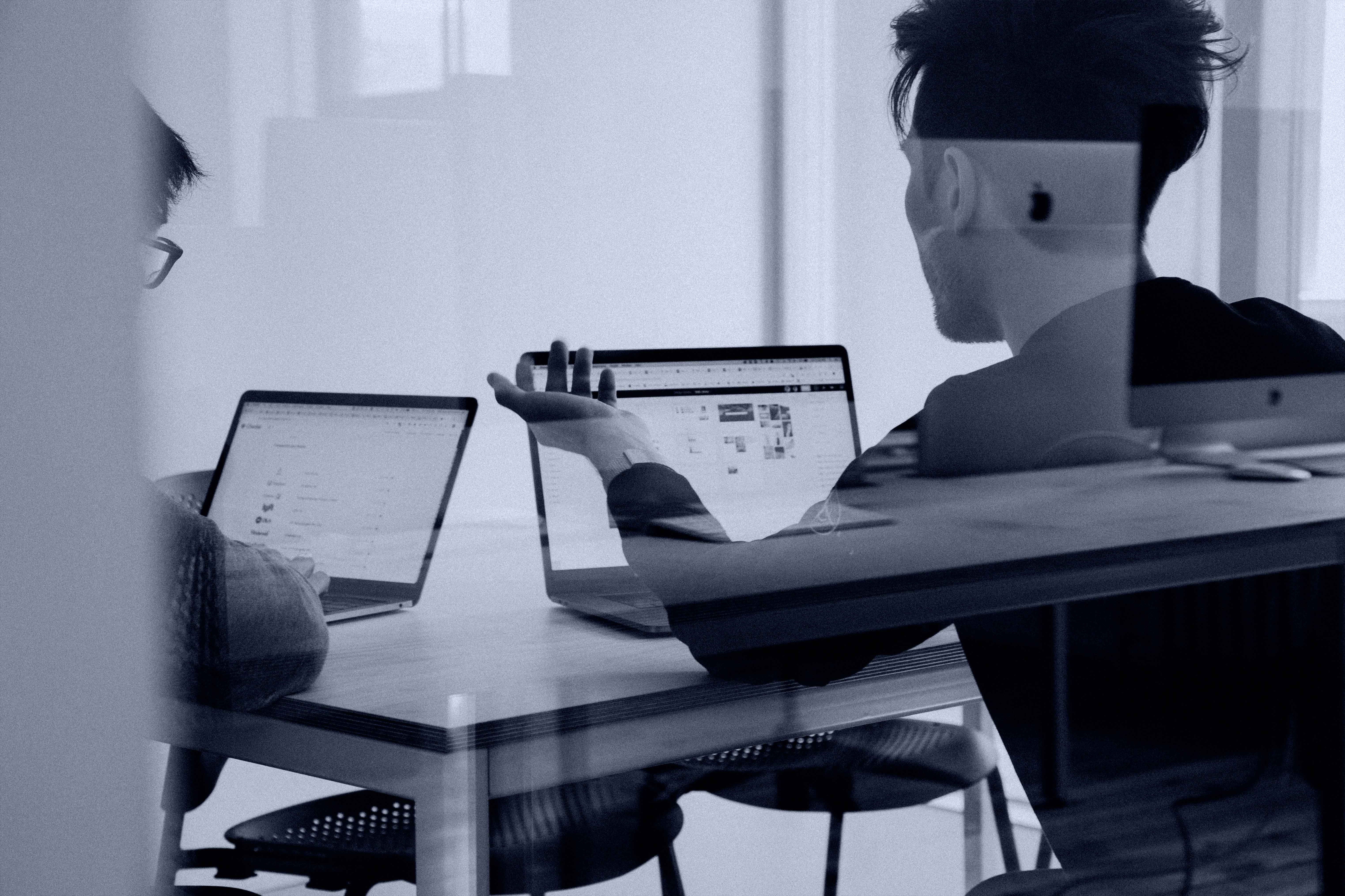 Photo through a reflective glass of two people in a meeting room, sitting in front of laptops