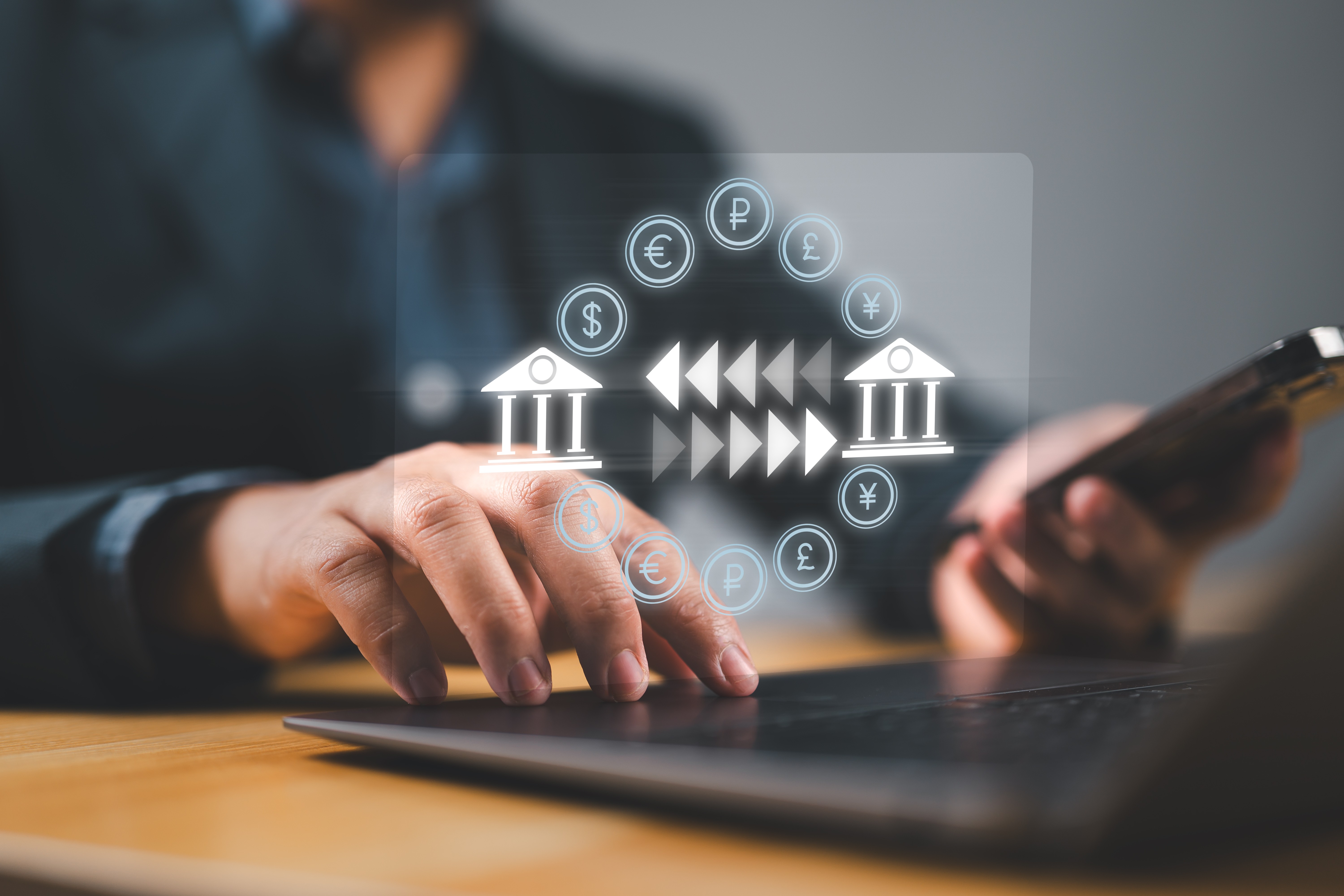 Person using a laptop and smartphone with digital icons representing international currency transfers between banks.