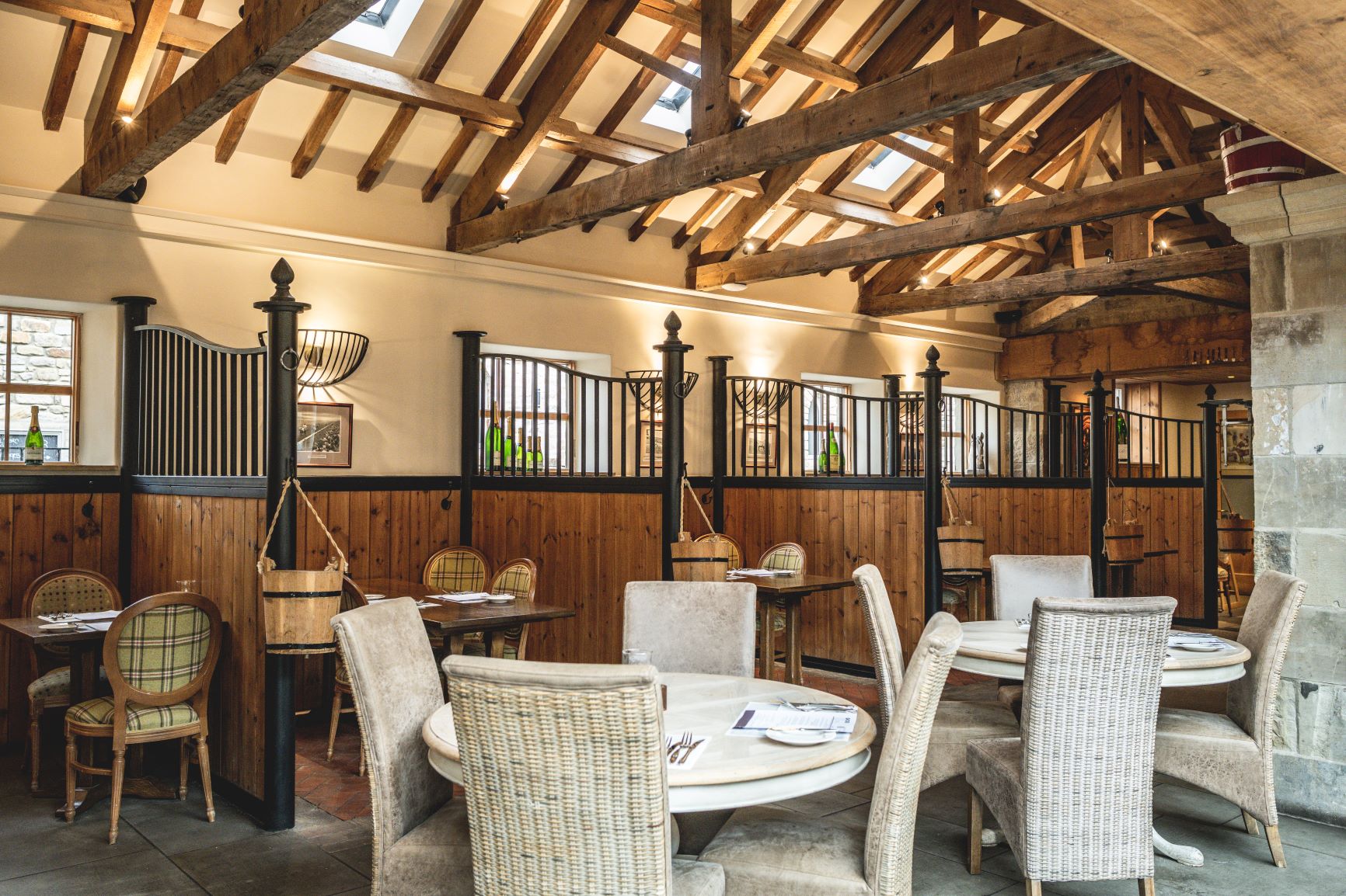 Elegant dining area in converted stable with exposed wooden beams, horse stalls and white wicker furniture arrangement