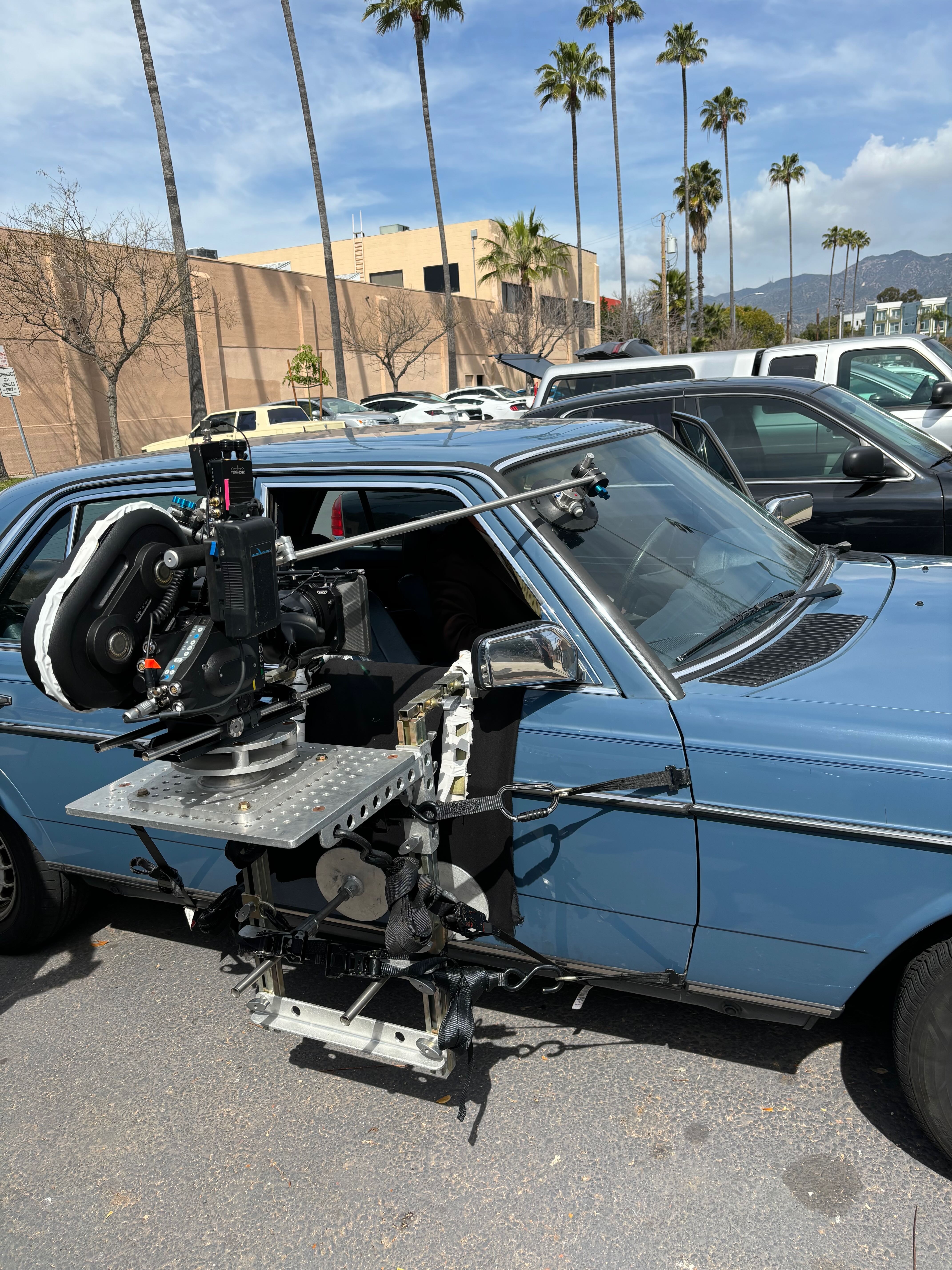 Cinema camera on a side platform beside a blue sedan, secured with straps and a windshield support rod
