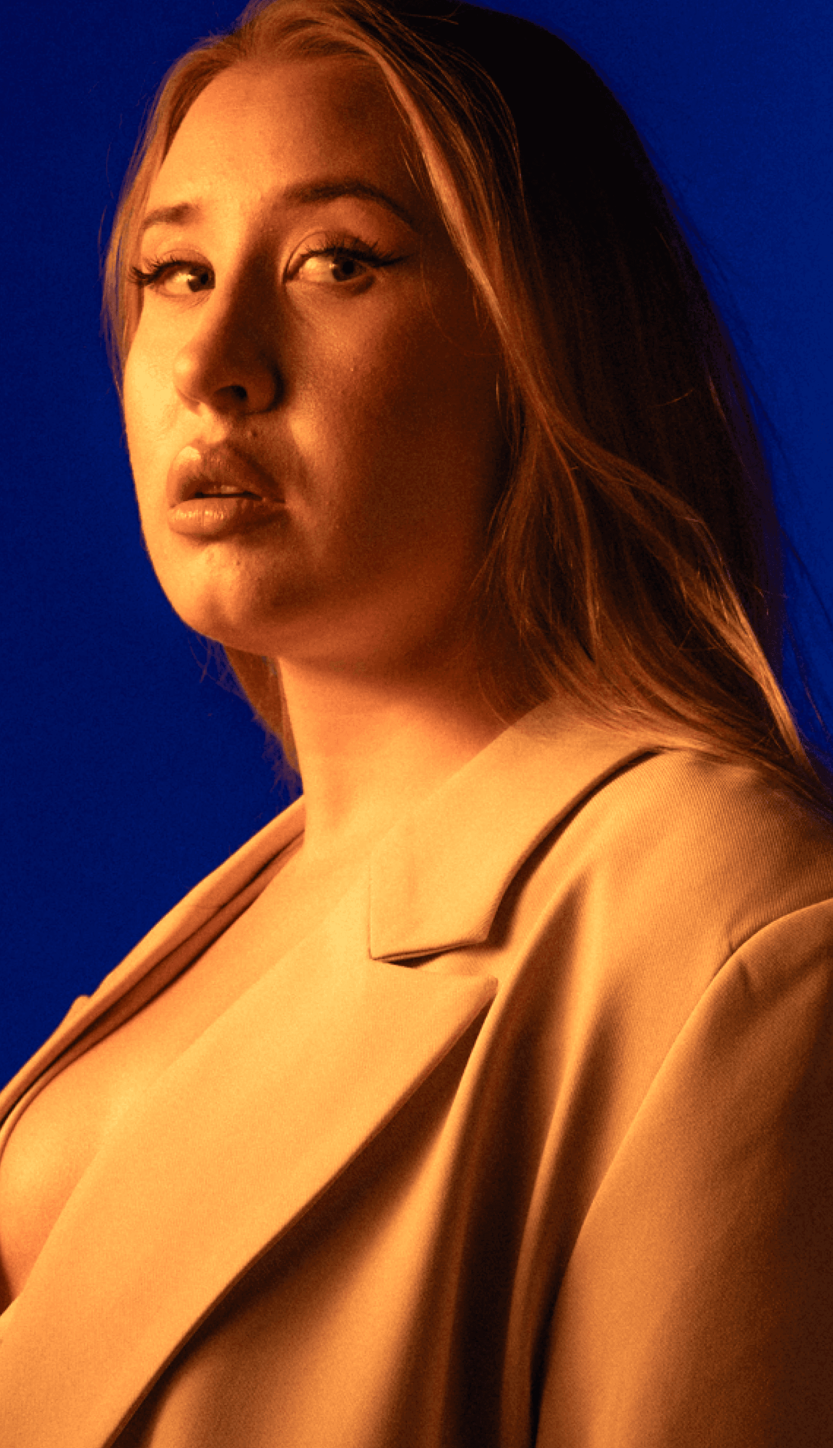 Dramatyczny portret członkini zespołu Electric Shapes – kobieta z długimi włosami w beżowej marynarce, oświetlona ciepłym, kontrastowym bursztynowym światłem na głębokim niebieskim tle