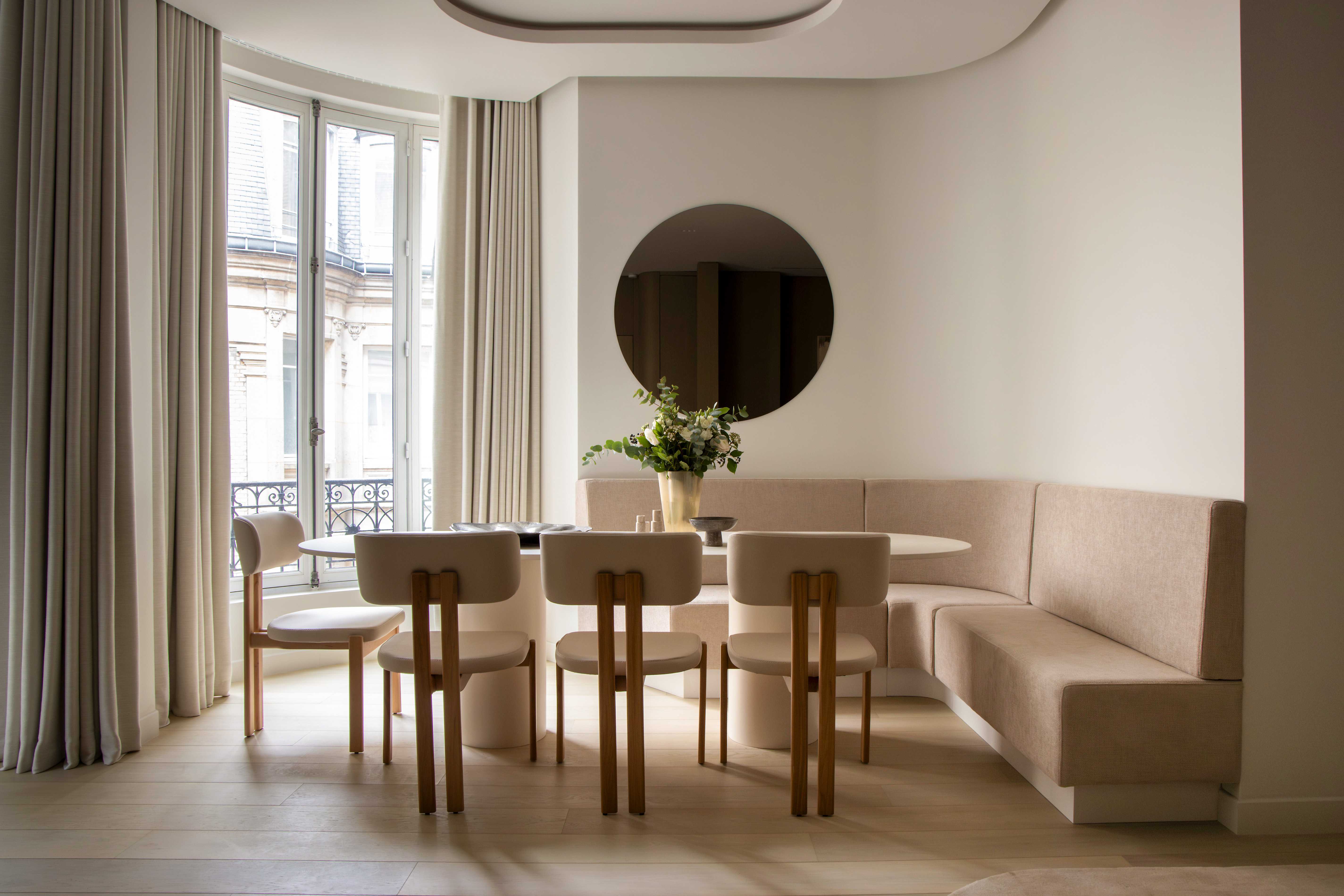 Coin repas avec banquette d'angle beige, chaises en bois, table ronde, miroir et vue sur balcon parisien