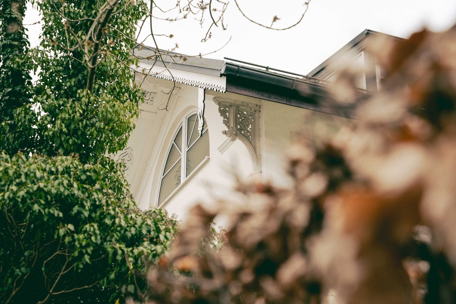 Nahaufnahme eines weißen Hauses mit einem Bogenfenster und dekorativen Verzierungen, umgeben von Bäumen und Blättern.