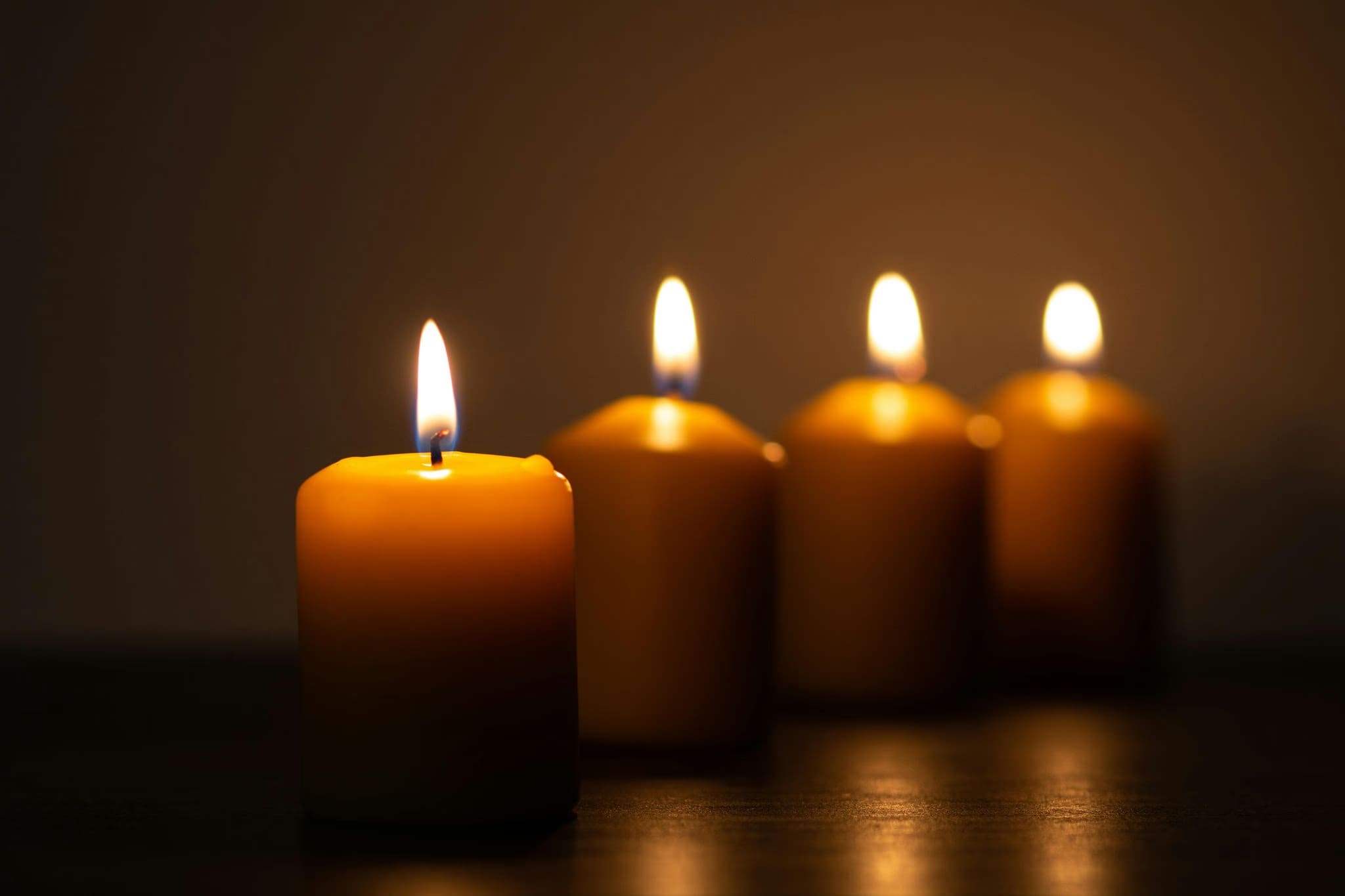 Four lit candles arranged in a row, with warm flames glowing in a dark, softly lit setting.