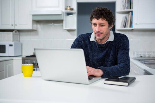 A man sits at his kitchen table with his laptop, notebook and yellow coffee mug and prepares to begin work