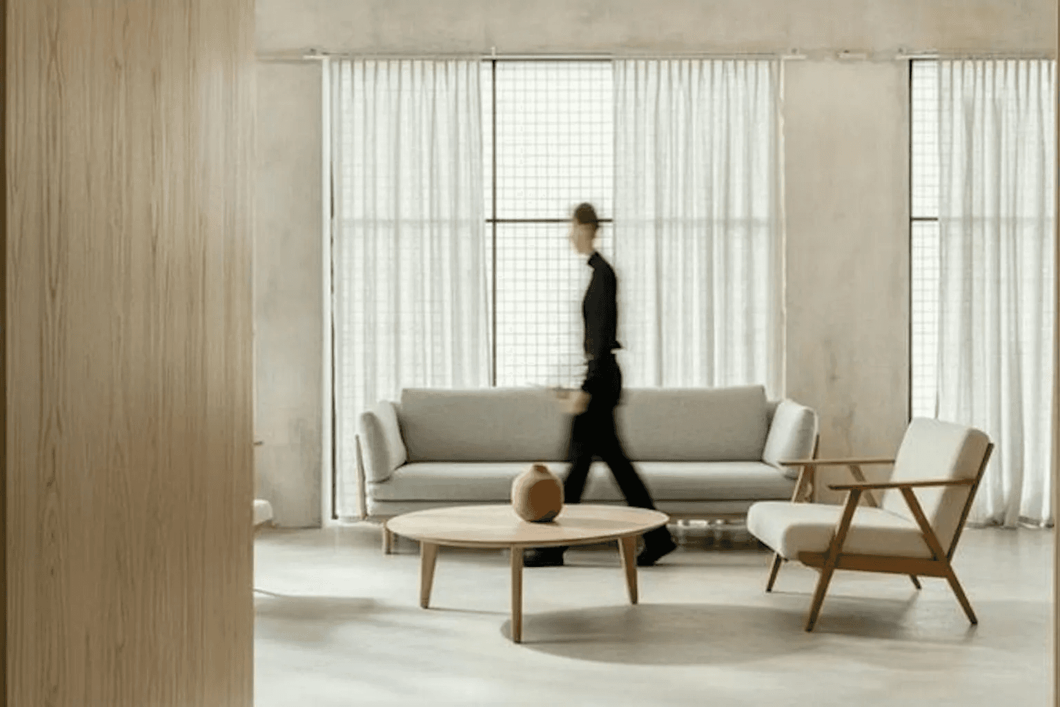 Minimalist living room with a grey sofa and wooden chairs as a blurred figure walks past.