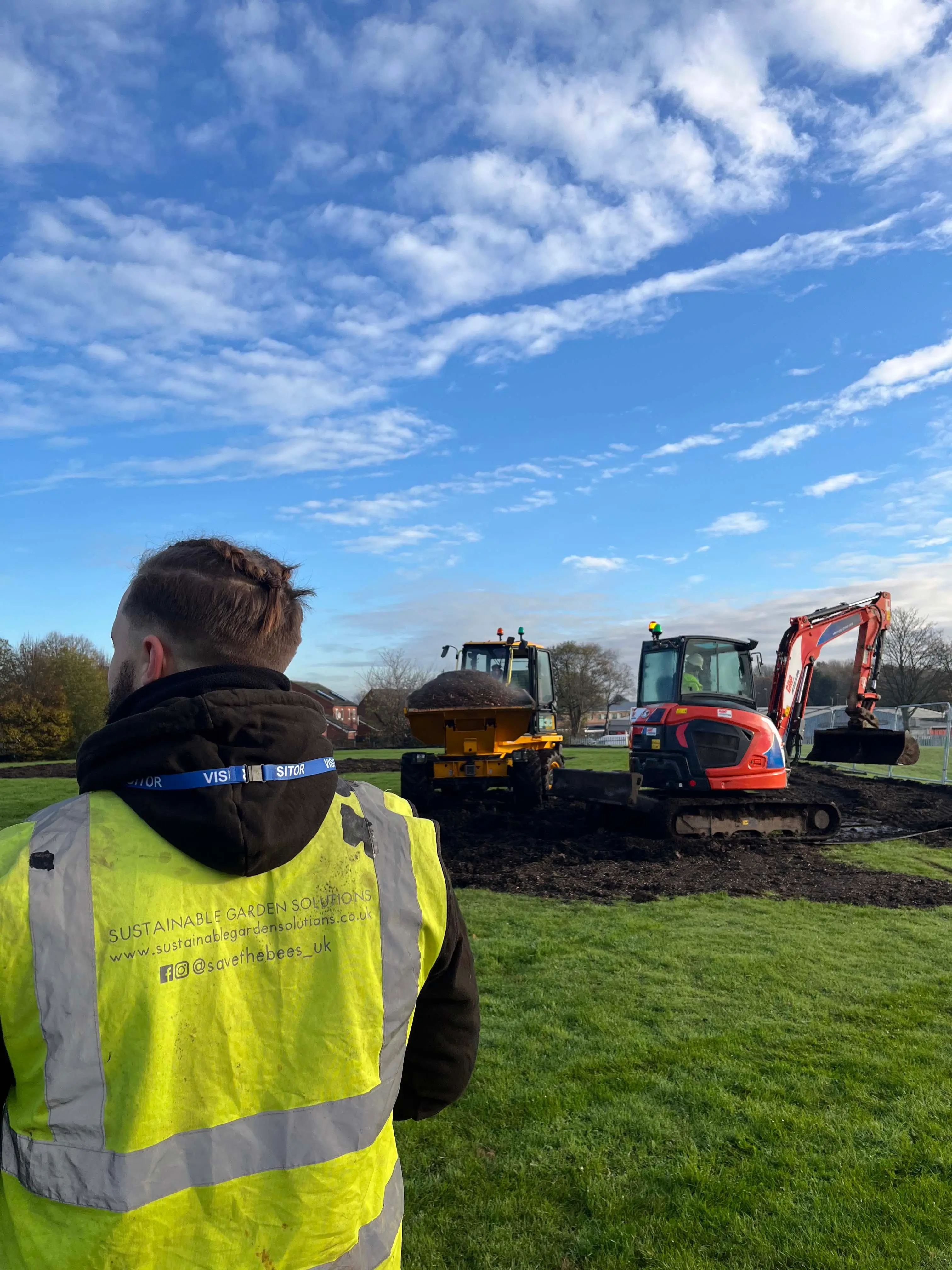 A person in a high-visibility vest stands facing away, watching machinery in a grassy field under a blue sky.