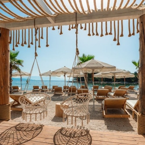 Strandscene med hengende kurvstoler, trelounger under stråparasoller og utsikt over havet.