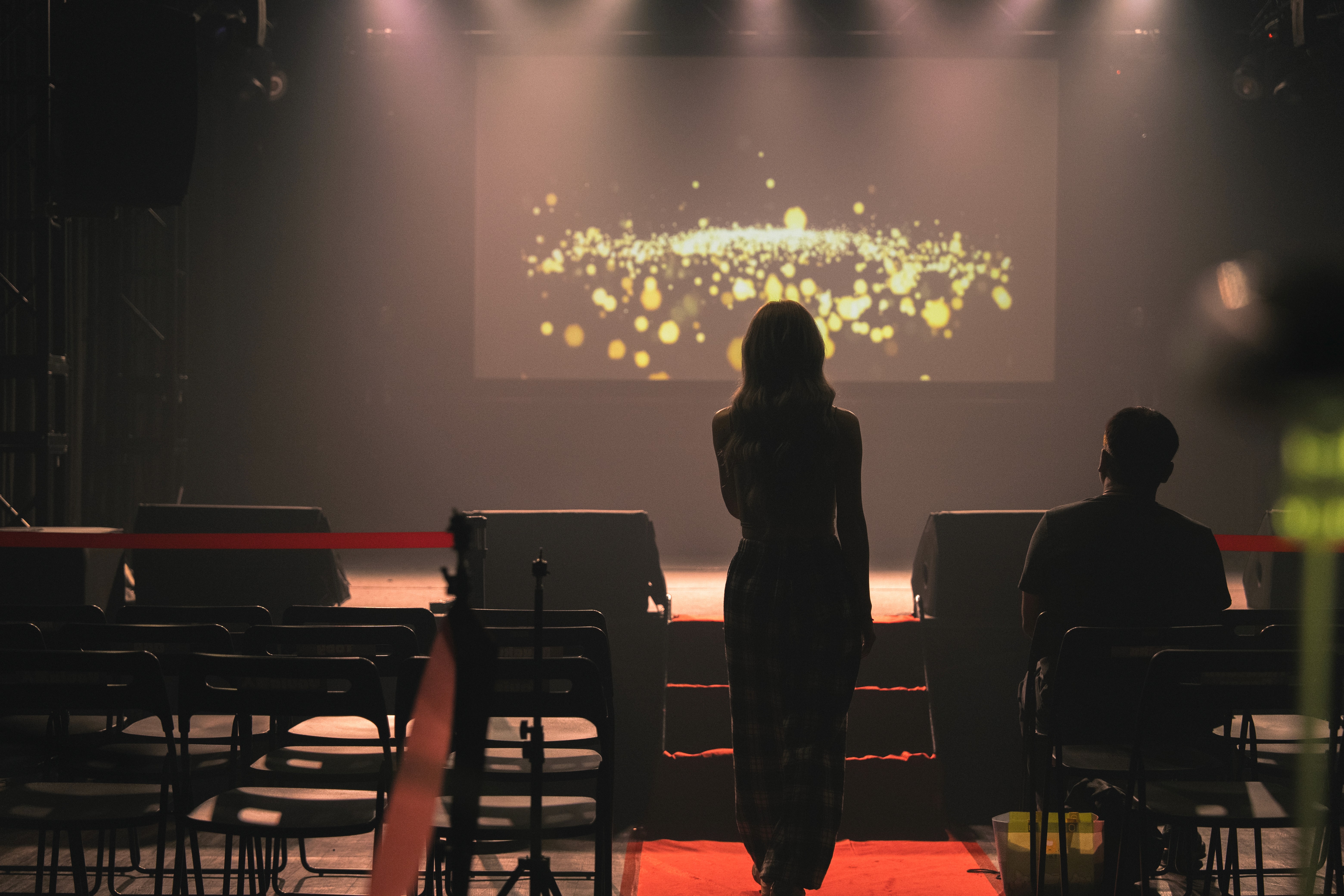 A person stands against a vibrant background of orange and yellow wavy patterns. They are wearing a dark coat, looking contemplative and thoughtful.