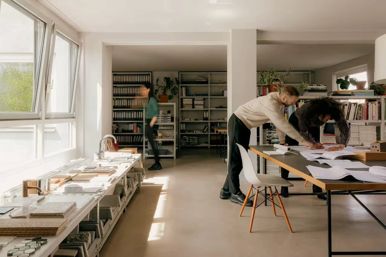 Two people work in a bright open-plan studio surrounded by shelves of binders and sample materials on tables.