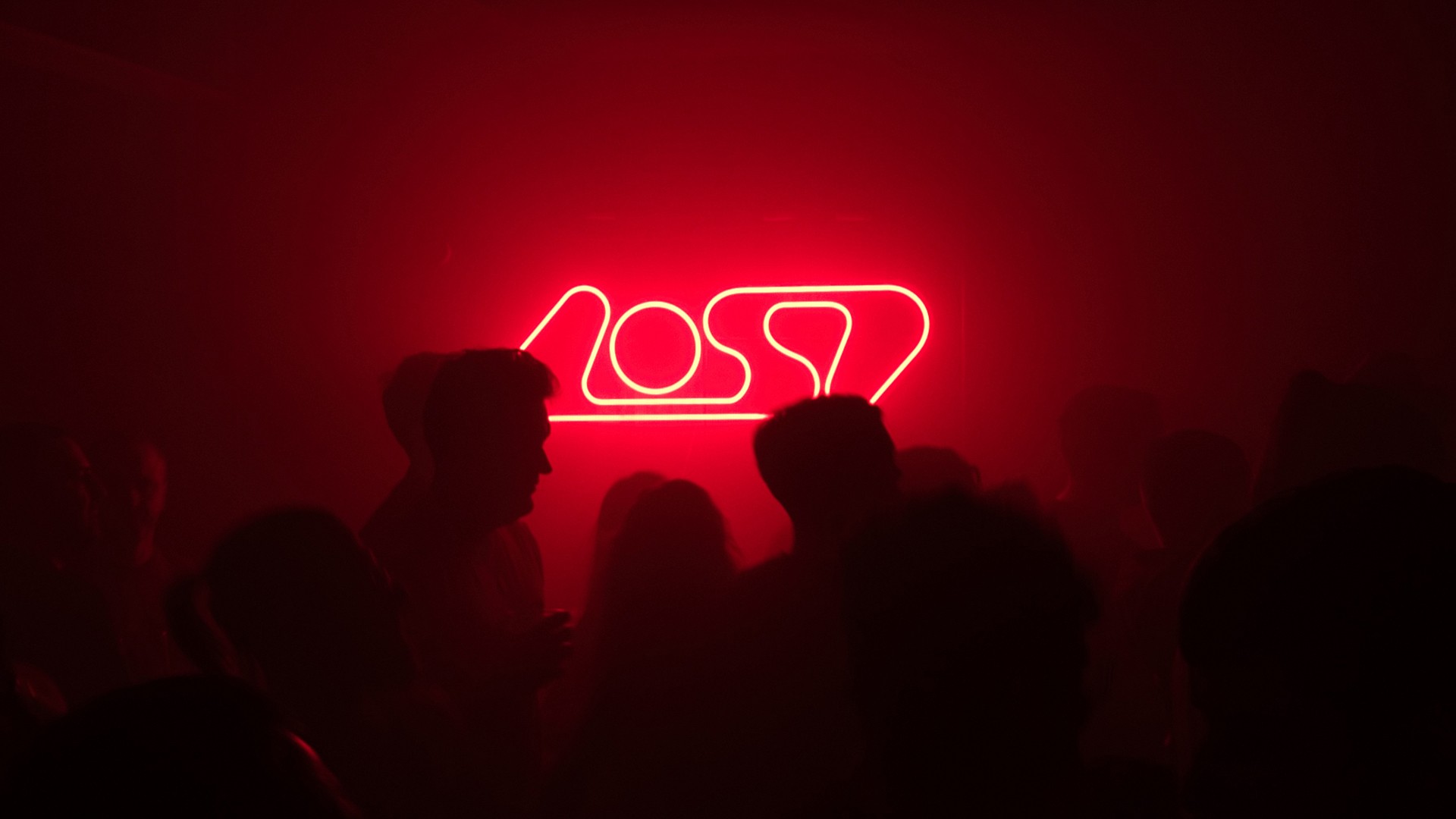 Red neon sign with the word “Lost” glowing above silhouettes of people in a dark room.
