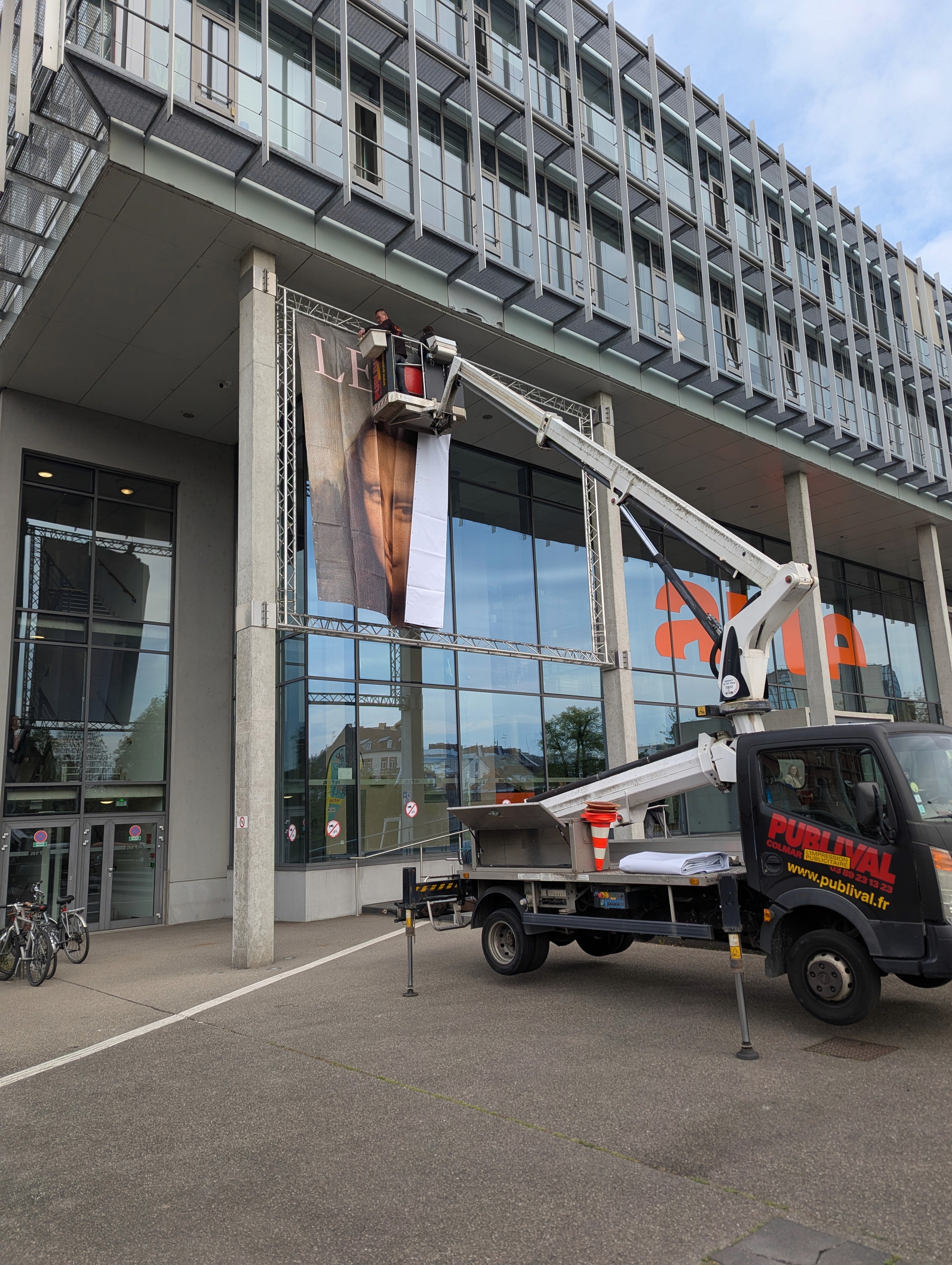 A large banner featuring a portrait of Mona Lisa is being hung on a modern building by workers in an aerial lift.