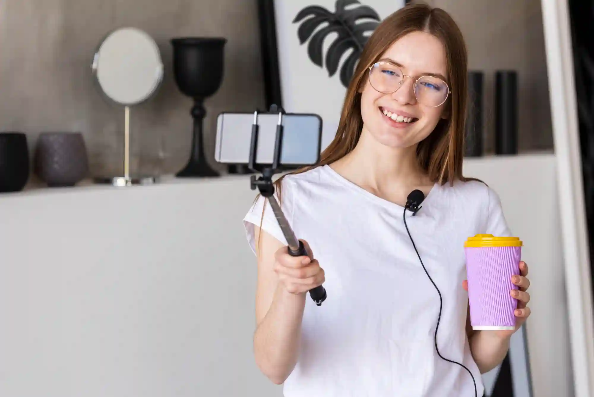 Smiling young woman filming video content with a smartphone on a stick, holding a pastel-colored thermal mug.