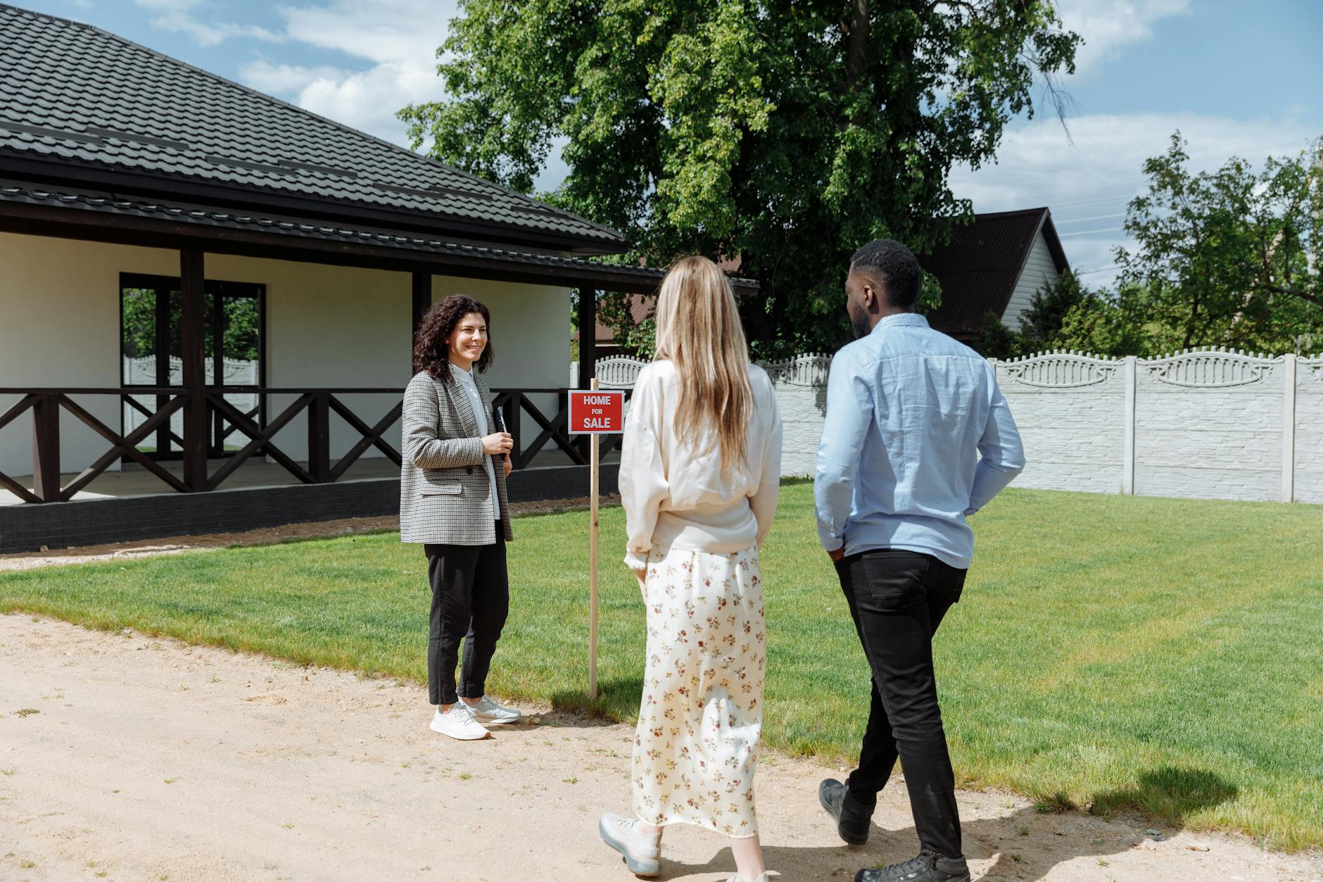 Two buyers walk toward a female estate agent standing beside a For Sale sign outside a white bungalow on a sunny day