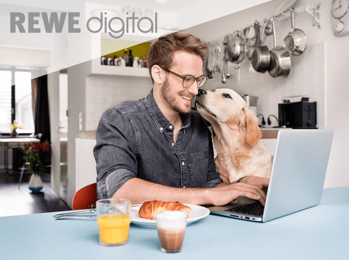 Ein Mann sitzt an seinem Tisch, Frühstückt und arbeitet am Laptop, neben ihm ist sein Hund der ihm gerade ein kleinen Kuss auf die Nase geben möchte, darauf befindet sich das Logo der Rewe Digital