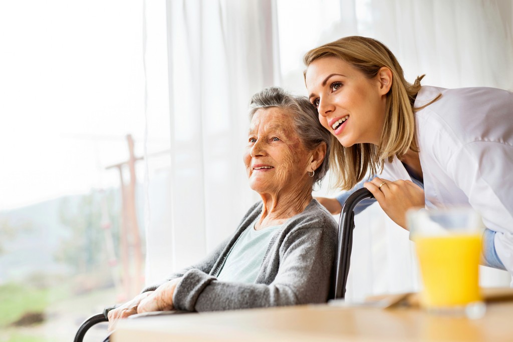 A cheerful caregiver assists an elderly woman in a wheelchair, both smiling and looking out the window, capturing a moment of companionship and attentive care in a bright, comfortable setting.