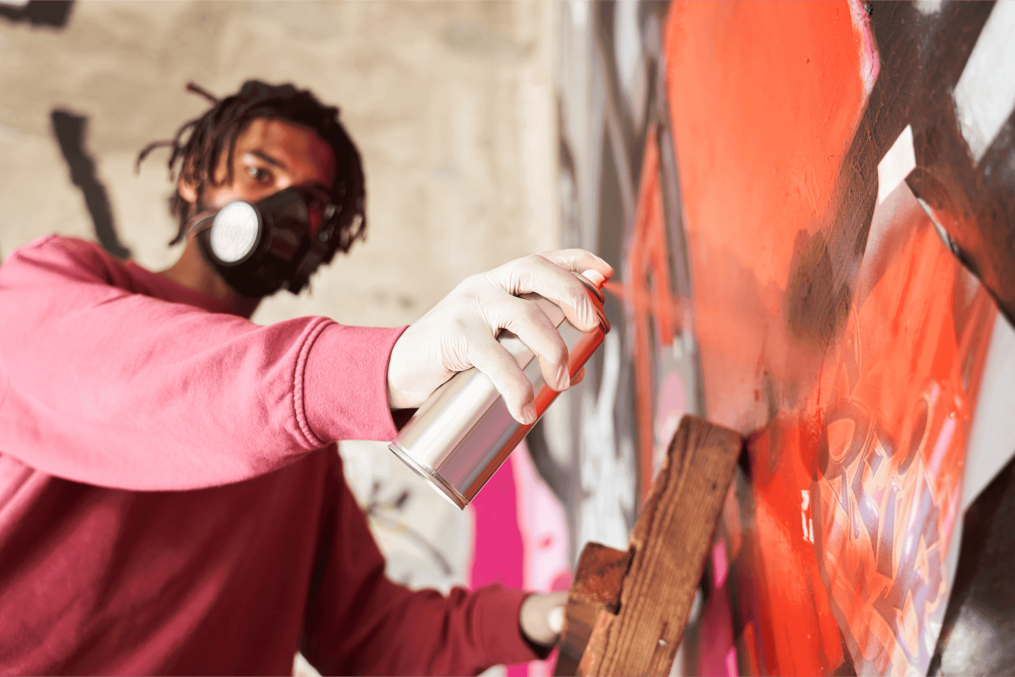 A person wearing a pink sweatshirt and a protective mask is using a spray paint can to create vibrant graffiti art on a large wall, with bold red and pink colors visible.