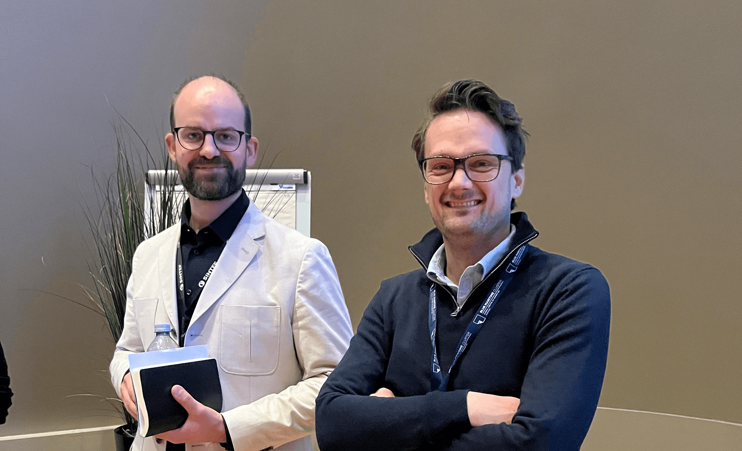 Two men stand smiling in a modern indoor setting, one wearing a white blazer and the other a dark sweater.