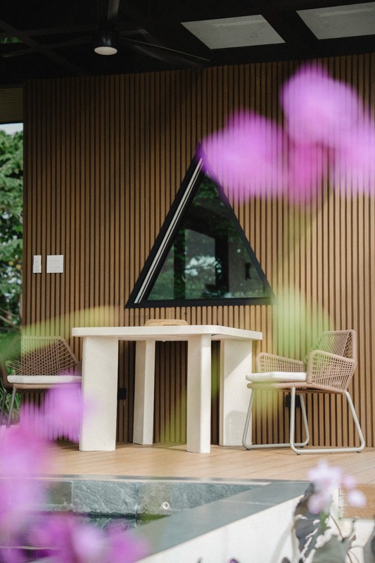 Outdoor dining nook with minimalist table, woven chairs, and triangular window set against vertical wood slats by the pool.