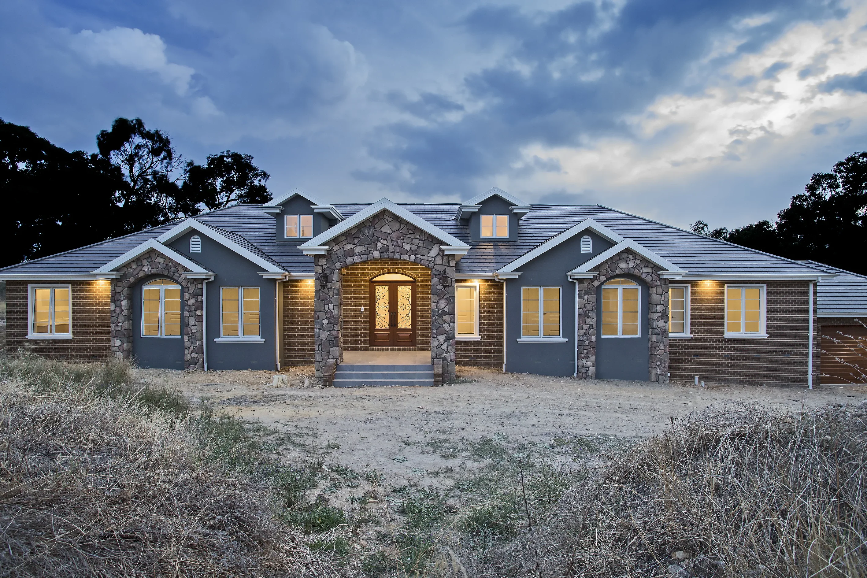 Front facade of Plenty Residence at dusk with grey-rendered and brick walls, natural stone arches, slate roof and a lit timber double-door entry