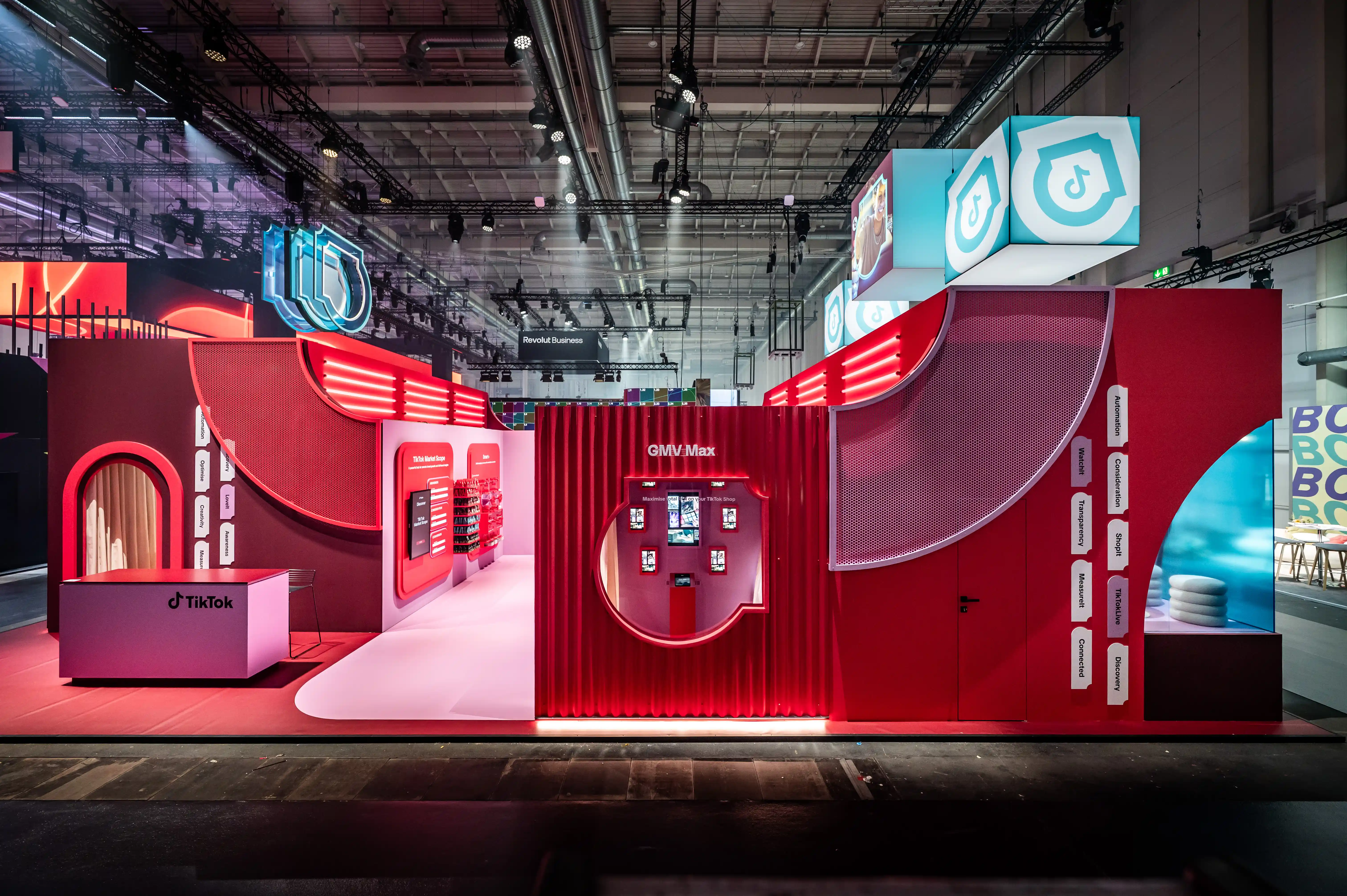 Long red and pink facade of the TikTok booth with GMV Max wall, counters and hanging shield cubes.