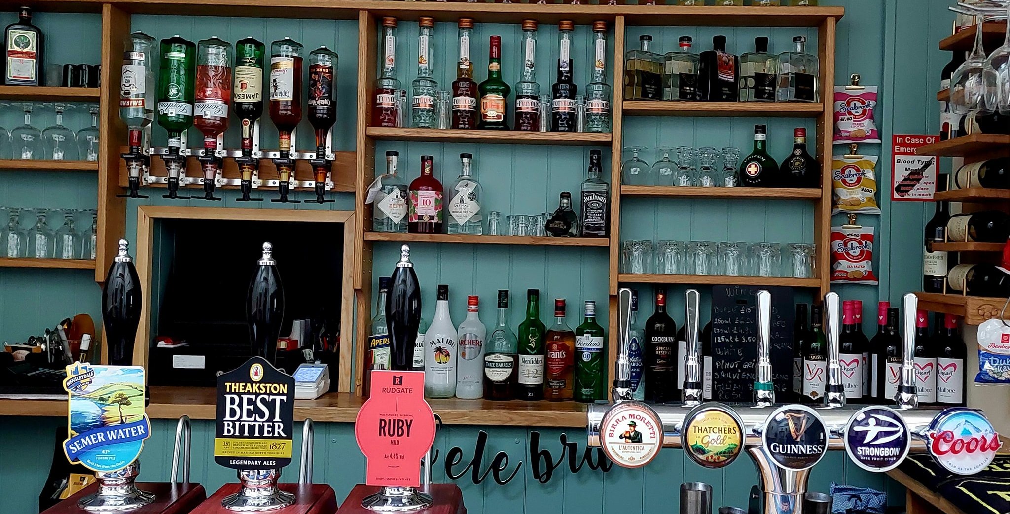 Bar shelving with wooden racks displaying various spirits and liquor bottles against a teal background. Beer taps and pump clips are visible in the foreground, showing a selection of draft beers available.